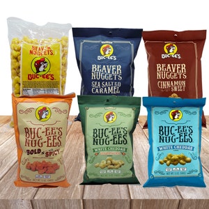 May include: Six bags of Buc-ee's Beaver Nuggets snack food. The bags are arranged in two rows of three. The flavors are: Bold 'n Spicy, White Cheddar Habanero, White Cheddar, Sea Salted Caramel, Cinnamon Sweet, and Original.