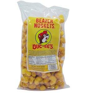 May include: A clear plastic bag filled with golden-yellow Beaver Nuggets. The bag has a yellow label with the text "BEAVER NUGGETS" and the Buc-ee's logo. The net weight is 13 oz (369g).