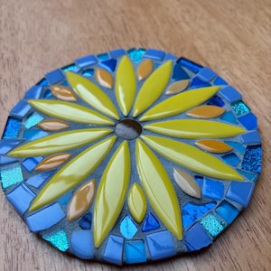 May include: A handcrafted mosaic coaster featuring a sunflower design. The coaster has a dark brown center with yellow petals, surrounded by a border of blue and turquoise mosaic tiles. The coaster is set on a wooden surface.
