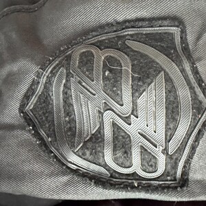 May include: A black fabric patch featuring a raised, moulded rubberised logo design in a shield shape. The emblem displays stylised, interconnected letters with a textured finish, stitched onto a dark, woven textile surface with visible seams.