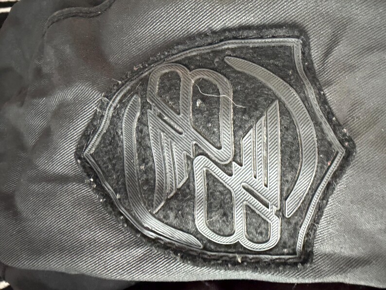 May include: A black fabric patch featuring a raised, moulded rubberised logo design in a shield shape. The emblem displays stylised, interconnected letters with a textured finish, stitched onto a dark, woven textile surface with visible seams.
