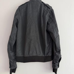 May include: A black jacket featuring a ribbed collar, ribbed cuffs, and a ribbed hem. The garment includes shoulder epaulets, long sleeves with buttoned cuffs, and vertical seam detailing along the back panel, presented on a hanger.