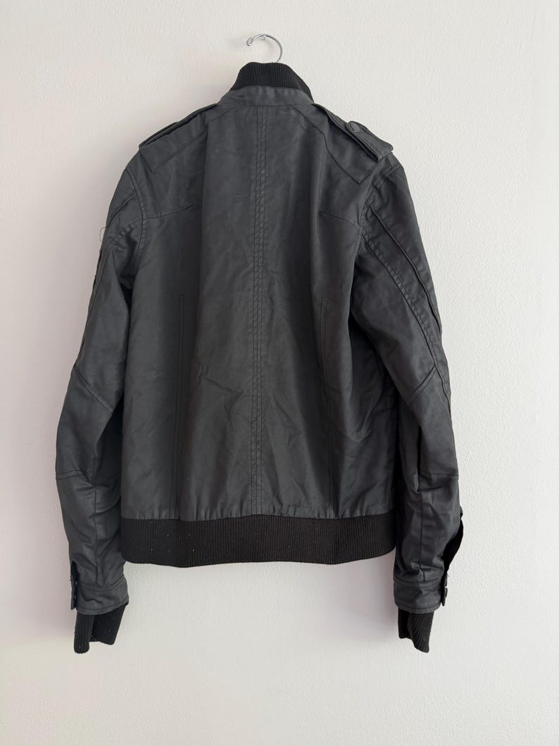May include: A black jacket featuring a ribbed collar, ribbed cuffs, and a ribbed hem. The garment includes shoulder epaulets, long sleeves with buttoned cuffs, and vertical seam detailing along the back panel, presented on a hanger.