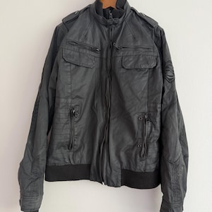 May include: A black jacket featuring a high ribbed collar, a central zip closure, and multiple zipped pockets on the chest and waist. The garment includes shoulder epaulettes, ribbed cuffs, and a circular emblem detail on the left sleeve.