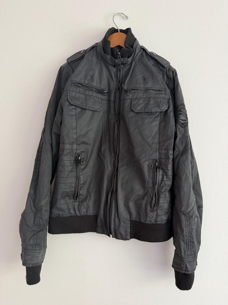 May include: A black jacket featuring a high ribbed collar, a central zip closure, and multiple zipped pockets on the chest and waist. The garment includes shoulder epaulettes, ribbed cuffs, and a circular emblem detail on the left sleeve.