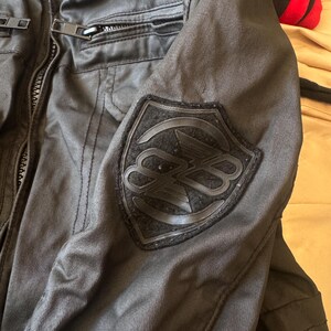 May include: A black jacket featuring a textured fabric construction with a front zip closure and a prominent shield-shaped patch on the sleeve. The patch displays a stylised circular logo with overlapping letters in a matching black tone.
