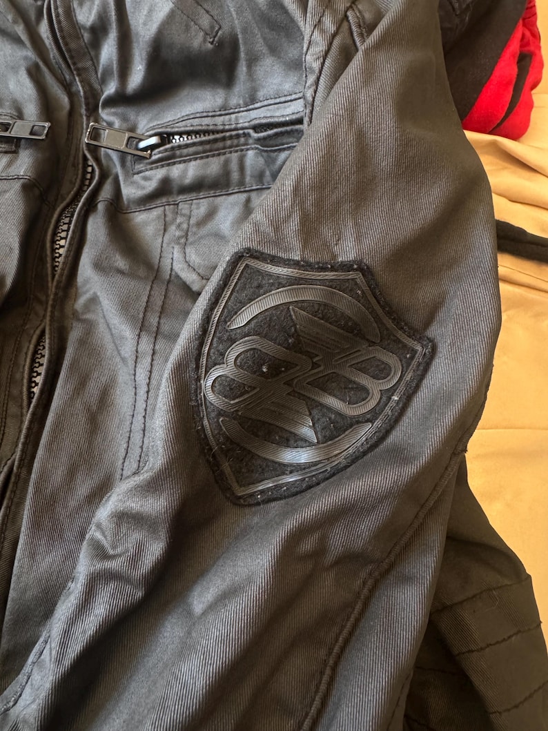 May include: A black jacket featuring a textured fabric construction with a front zip closure and a prominent shield-shaped patch on the sleeve. The patch displays a stylised circular logo with overlapping letters in a matching black tone.