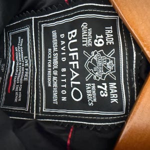 May include: A black garment interior label featuring white embroidered text that reads Buffalo David Bitton, Trade Mark 1973, Live Free, and other branding details. The rectangular patch has a decorative scalloped edge and is stitched onto black fabric.