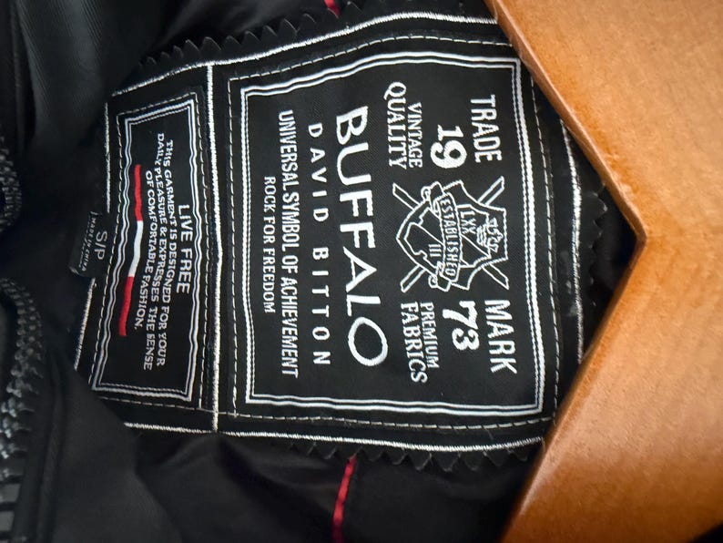 May include: A black garment interior label featuring white embroidered text that reads Buffalo David Bitton, Trade Mark 1973, Live Free, and other branding details. The rectangular patch has a decorative scalloped edge and is stitched onto black fabric.