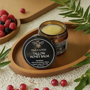 May include: A black jar with a gold label that reads "TERRA LOTUS TALLOW HONEY BALM" and lists the ingredients: 100% Grass-Fed Beef Tallow, Organic Italian Olive Oil, Raw Honey. The jar is sitting on a wooden surface with red berries scattered around it.