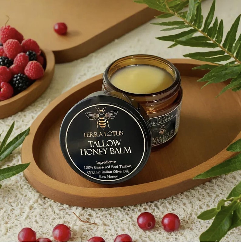 May include: A black jar with a gold label that reads "TERRA LOTUS TALLOW HONEY BALM" and lists the ingredients: 100% Grass-Fed Beef Tallow, Organic Italian Olive Oil, Raw Honey. The jar is sitting on a wooden surface with red berries scattered around it.