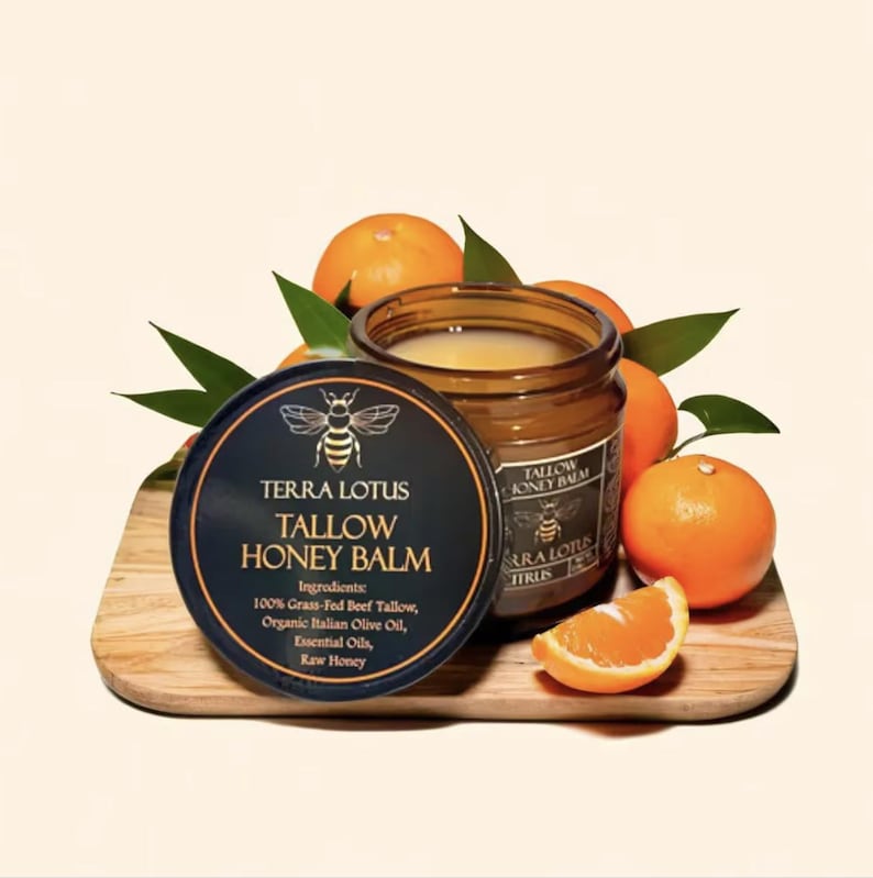 May include: A glass jar of Terra Lotus Tallow Honey Balm with a black lid featuring a bee logo and the product name. The jar is sitting on a wooden surface with orange slices and green leaves.