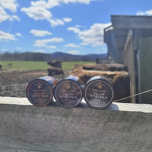 May include: Three small black jars with gold lids and labels that read "Terra Lotus Tallow Honey Balm". The jars are sitting on a wooden surface.