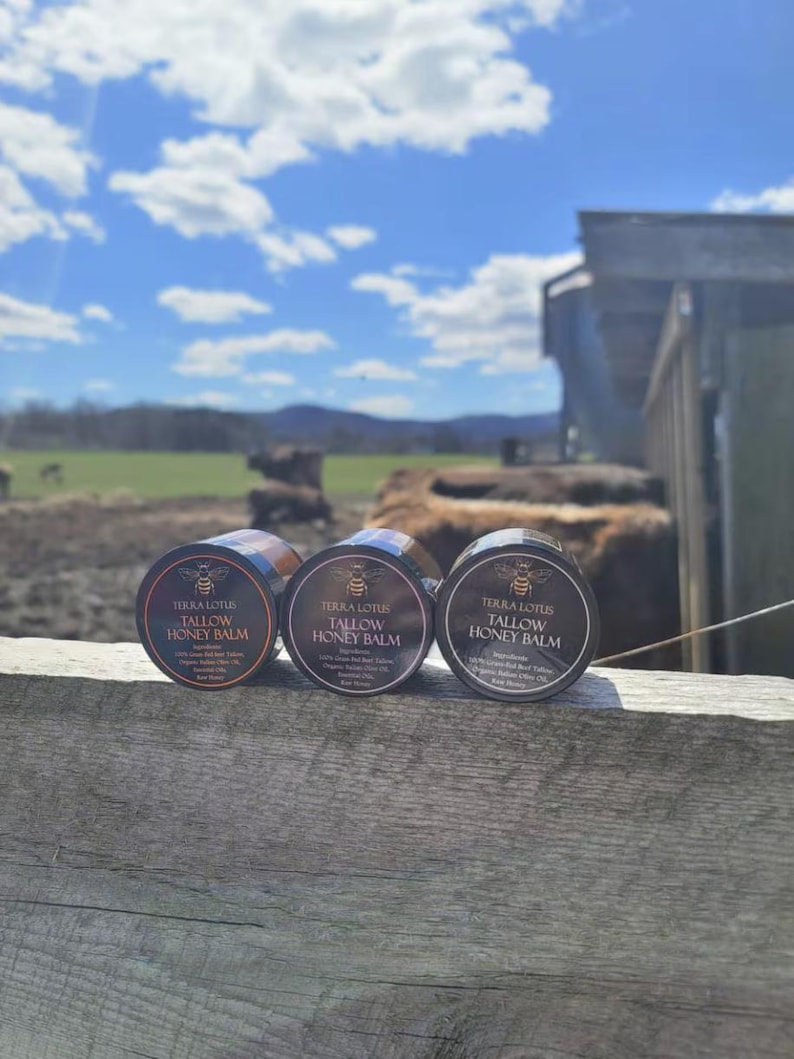 May include: Three small black jars with gold lids and labels that read "Terra Lotus Tallow Honey Balm". The jars are sitting on a wooden surface.