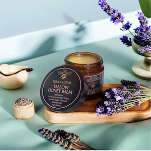 May include: A brown glass jar with a black lid containing a yellow balm. The jar is labeled "Terra Lotus Tallow Honey Balm" and lists the ingredients: 100% Grass-Fed Beef Tallow, Organic Italian Olive Oil, Essential Oils, Raw Honey. The jar is sitting on a wooden surface with lavender flowers.