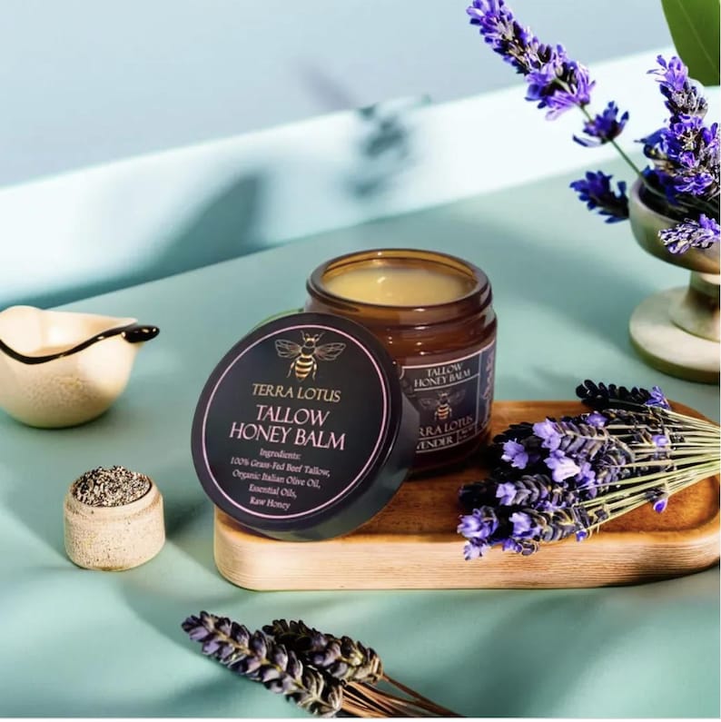 May include: A brown glass jar with a black lid containing a yellow balm. The jar is labeled "Terra Lotus Tallow Honey Balm" and lists the ingredients: 100% Grass-Fed Beef Tallow, Organic Italian Olive Oil, Essential Oils, Raw Honey. The jar is sitting on a wooden surface with lavender flowers.