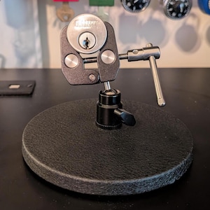 May include: A black metal lock vice mounted on a circular textured base. The vice features a silver-coloured cylinder lock held by two adjustable arms with a side-mounted tightening handle. The device is designed to secure locks for inspection.