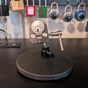 May include: A black and silver lock pick tool mounted on a round, textured black base. The tool has a silver handle and is designed to hold a lock. Various locks hang in the background.