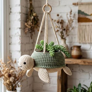 May include: A hanging crochet planter shaped like a turtle with a cream-colored head and flippers, and a sage green shell that holds a green succulent plant. The planter is suspended by four thick cream-colored cords attached to a wooden ring.