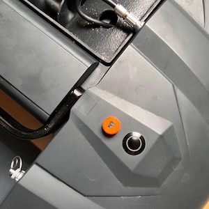 Op de afbeelding: Een donkergrijs mechanisch apparaat met een zwart vijzelmechanisme, een zwarte kabel met een metalen connector, een kleine ronde oranje knop met een zwarte letter F en een grotere ronde zwarte drukknop met een zilverkleurige metalen rand.