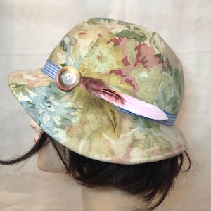 Può includere: Un cappello a secchiello con motivo floreale nei toni del verde, rosa e crema. Il cappello è decorato con un nastro a quadretti blu e bianco, un singolo bottone rotondo e una piuma lunga e sottile nei toni del rosa e del verde oliva.
