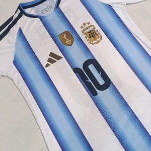 May include: A white and light blue striped football shirt featuring the number 10 in dark blue, the AFA crest with three gold stars, a gold FIFA 2022 World Cup patch, a black logo, and dark blue shoulder stripes on a short-sleeved jersey.