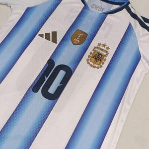 May include: A white and light blue striped football shirt featuring the number 10 in dark blue on the front. The jersey includes a gold FIFA 2022 World Cup patch, the AFA crest with three gold stars, and a black Adidas logo on the chest area.