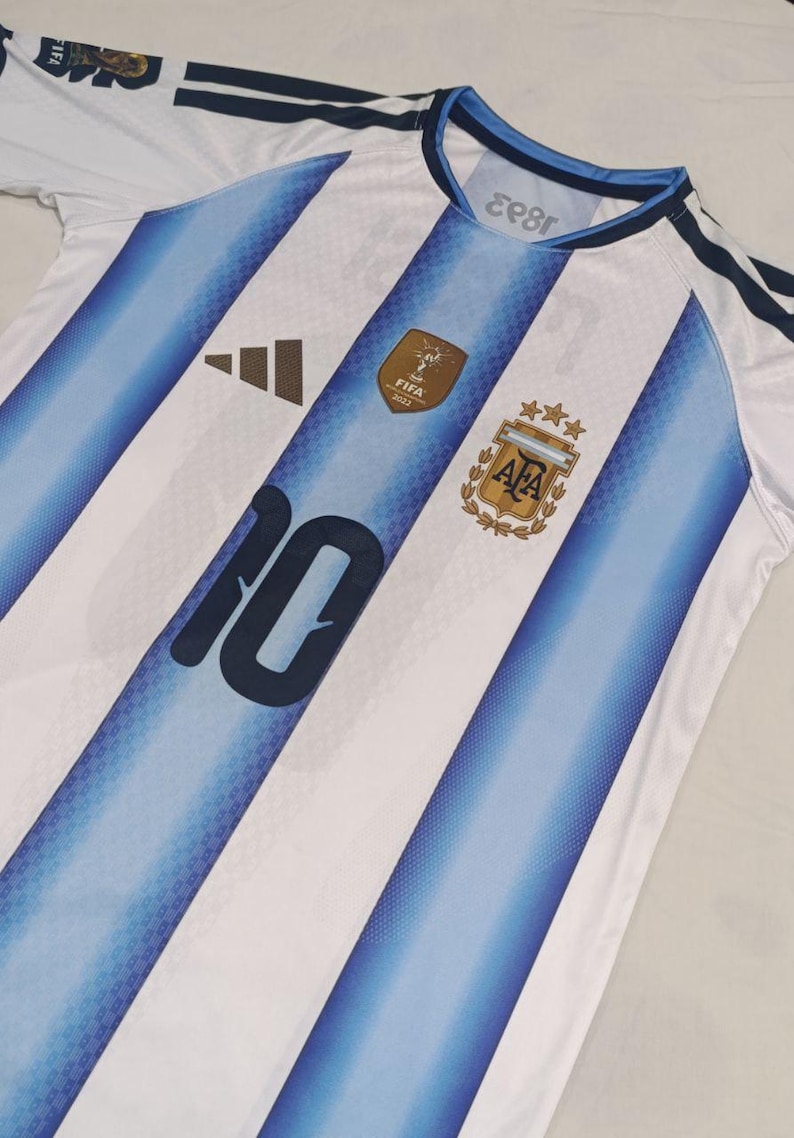 May include: A white and light blue striped football shirt featuring the number 10 in dark blue on the front. The jersey includes a gold FIFA 2022 World Cup patch, the AFA crest with three gold stars, and a black Adidas logo on the chest area.