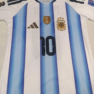 May include: A white and light blue striped football shirt featuring the number 10 in dark blue on the front. The jersey includes a gold FIFA 2022 World Cup patch, the Argentina national team crest with three gold stars, and a black Adidas logo.