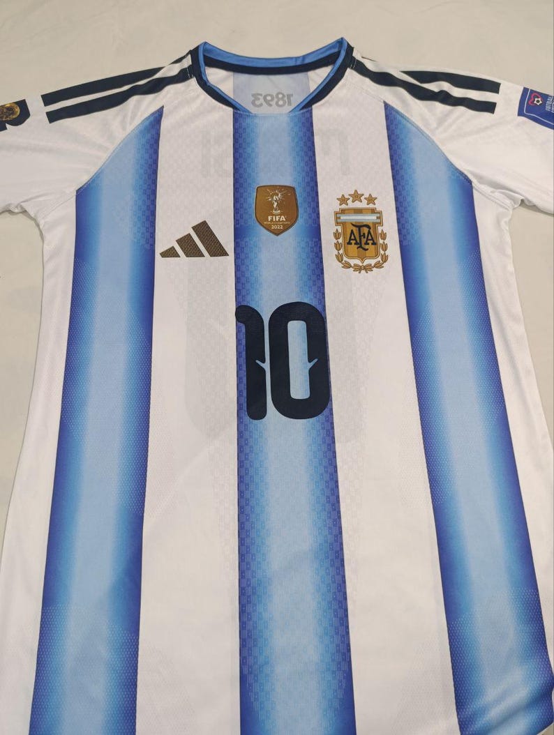 May include: A white and light blue striped football shirt featuring the number 10 in dark blue on the front. The jersey includes a gold FIFA 2022 World Cup patch, the Argentina national team crest with three gold stars, and a black Adidas logo.