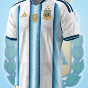 May include: A white and light blue striped football shirt featuring the Argentina national team crest with three gold stars, a gold FIFA World Cup winner badge, and a gold Adidas logo on the chest. The shirt has short sleeves and a V-neck collar.