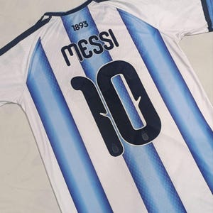 May include: A white and light blue striped sports jersey featuring the name MESSI and the number 10 printed in dark blue on the back. The text 1893 is printed above the name. The jersey has dark blue trim on the collar and shoulder sleeves.