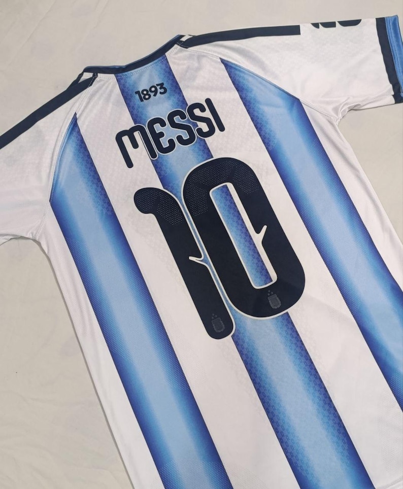 May include: A white and light blue striped sports jersey featuring the name MESSI and the number 10 printed in dark blue on the back. The text 1893 is printed above the name. The jersey has dark blue trim on the collar and shoulder sleeves.