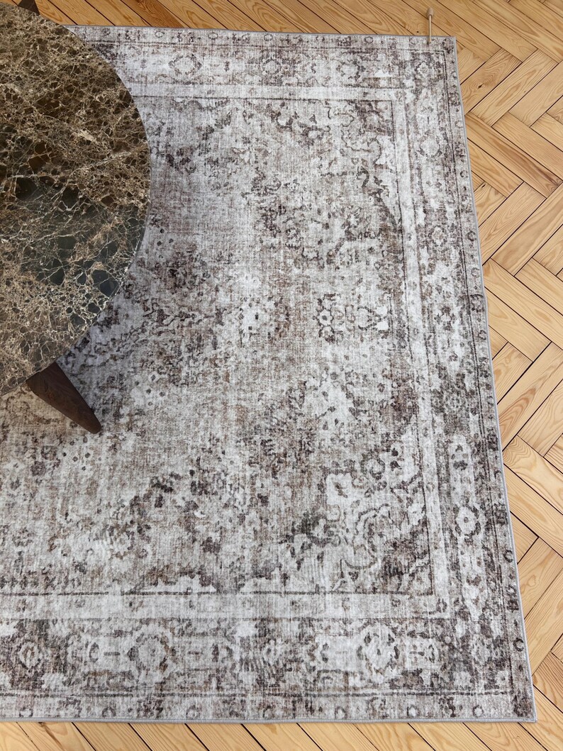 May include: A rectangular area rug with a distressed, vintage-inspired design. The rug features a central field of beige and brown tones with an intricate floral pattern. A dark brown border frames the design. A round marble table is partially visible.