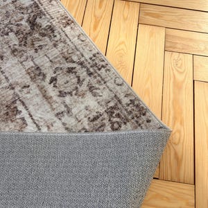 May include: A corner of a beige and brown patterned rug with a gray backing, placed on a light-colored wooden floor. The rug's design features a distressed, vintage aesthetic, with floral and geometric elements.