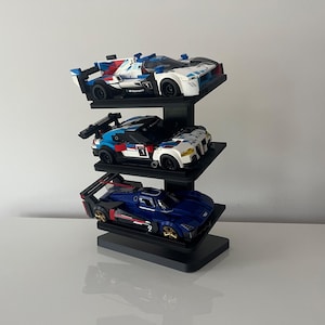 May include: A black three-tiered display stand holding three model race cars. The top car is white, blue, and red with the number 1. The middle car is white, blue, and red with the number 1. The bottom car is dark blue with gold wheels and the number 9.