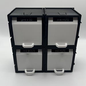 May include: A black and white storage organizer with four drawers. Each drawer is labeled with text: "M4 x 50", "M4 NUTS", "M5 BOLTS", and "M5 NUTS". The drawers have white fronts and white handles.
