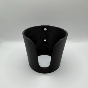 May include: A black, cylindrical wall-mounted holder featuring a textured exterior surface. The holder has a large, U-shaped cutout on the front and two vertically aligned circular mounting holes on the back panel for secure attachment to a flat surface.