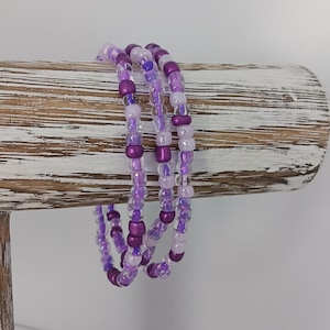May include: Three purple and white beaded bracelets stacked on a wooden display stand. The bracelets are made with small seed beads in various shades of purple and white.