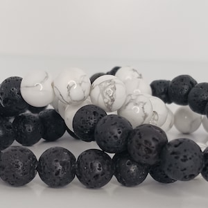 May include: Close-up of two beaded bracelets. One bracelet features black lava rock beads, while the other alternates between white marble-like beads and black lava rock beads. The beads are round and smooth.