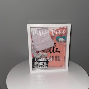 May include: A white shadow box with a pink and white striped onesie, a pink onesie with the text "hello" and "new here", a pacifier, and a white card with the text "Oaklyn Grace", "08/17/2024", "LBS 902", and "9:07PM".