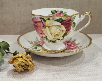 May include: A white teacup and saucer set with a floral rose design in shades of pink, yellow, and red. The cup and saucer have gold trim. A dried yellow rose is next to the set. The teacup is approximately 3 inches tall.