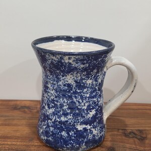 May include: A ceramic mug featuring a speckled blue and white glaze pattern. The mug has a flared rim, a cylindrical body with subtle horizontal ridges, and a sturdy, curved handle. The interior and the handle are finished in a solid off-white.