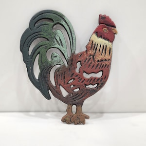May include: A cast iron rooster trivet featuring a cutout design. The rooster is painted with a red head, a brown and tan body, and a dark green tail. The metal surface has a textured, weathered finish with visible paint distressing throughout.