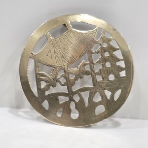 May include: A circular brass trivet featuring an intricate cutout design of a pagoda, a pavilion, and flying birds. The metallic surface has a polished gold tone with detailed etchings depicting architectural structures and natural elements.