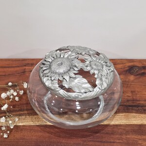 May include: A clear glass bowl featuring a removable silver-toned metal lid. The lid is decorated with a relief design of three sunflowers with textured centres and detailed leaves, creating a circular floral pattern across the top surface.