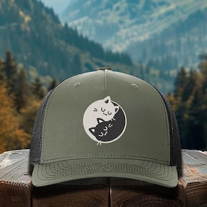 May include: A green and black trucker hat with a white and black yin yang symbol featuring two cats. The cats are facing each other and have closed eyes.