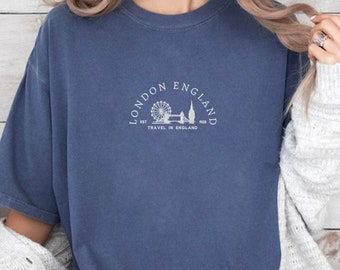 May include: A denim blue t-shirt with a white graphic of the London Eye and the text "LONDON ENGLAND EST 1999 TRAVEL IN ENGLAND".
