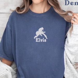 May include: Denim blue t-shirt featuring a white silhouette of a musician playing a guitar, with the name "Elvis" below. The shirt has short sleeves and a crew neck. The word "Denim" is printed in the upper right corner.