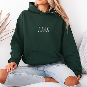 May include: Dark green hoodie with a front pocket and a drawstring hood. The hoodie features an embroidered design of four figures walking in a line. The model is wearing light blue jeans. The hoodie is made of a soft material.